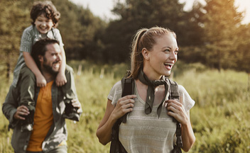 Eine Frau mit einem Rucksack lächelt, während sie in einem Feld läuft. Ein Mann trägt einen Jungen auf den Schultern. Sie sind in einer natürlichen Umgebung mit Bäumen im Hintergrund.