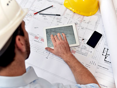 Ein Bauarbeiter mit einem Helm, der auf einem Tablet arbeitet, umgeben von Bauplänen, einem Smartphone und Bleistiften.