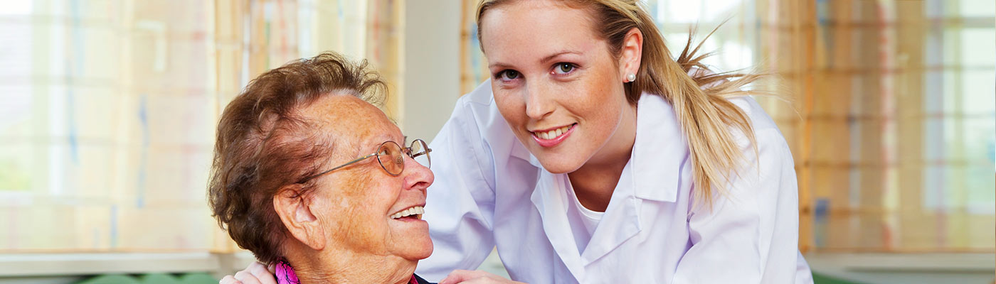 Eine ältere Frau lächelt und schaut mit einer Pflegekraft, die ihr freundlich gegenübersteht. Beide strahlen Freude aus.