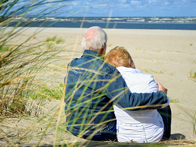 Ein älteres Paar sitzt am Strand und umarmt sich, im Hintergrund ist das Meer und der Himmel zu sehen.