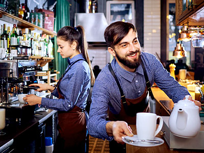 Zwei Baristas in einem Café, einer serviert eine Tasse, die andere bereitet Kaffee zu.