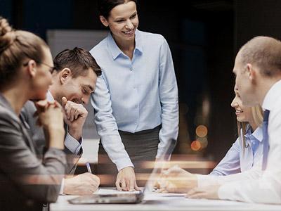 Ein Team von fünf Personen in einem Besprechungsraum, wobei eine Frau an einem Tisch steht und Informationen präsentiert, während die anderen Teilnehmer Notizen machen.