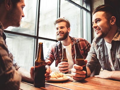 Drei Männer sitzen an einem Tisch, trinken Bier und unterhalten sich. Am Tisch steht ein Teller mit Snacks.