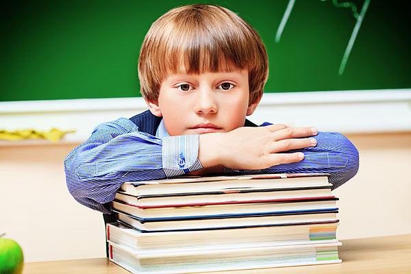 Ein Junge mit blonden Haaren lehnt auf einem Stapel Bücher und schaut nachdenklich. Im Hintergrund ist eine grüne Tafel zu sehen.