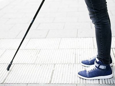 Ein Bein einer Person mit einem Gehstock steht auf einem Gehweg. Die Person trägt blaue Sneakers und schwarze Jeans.